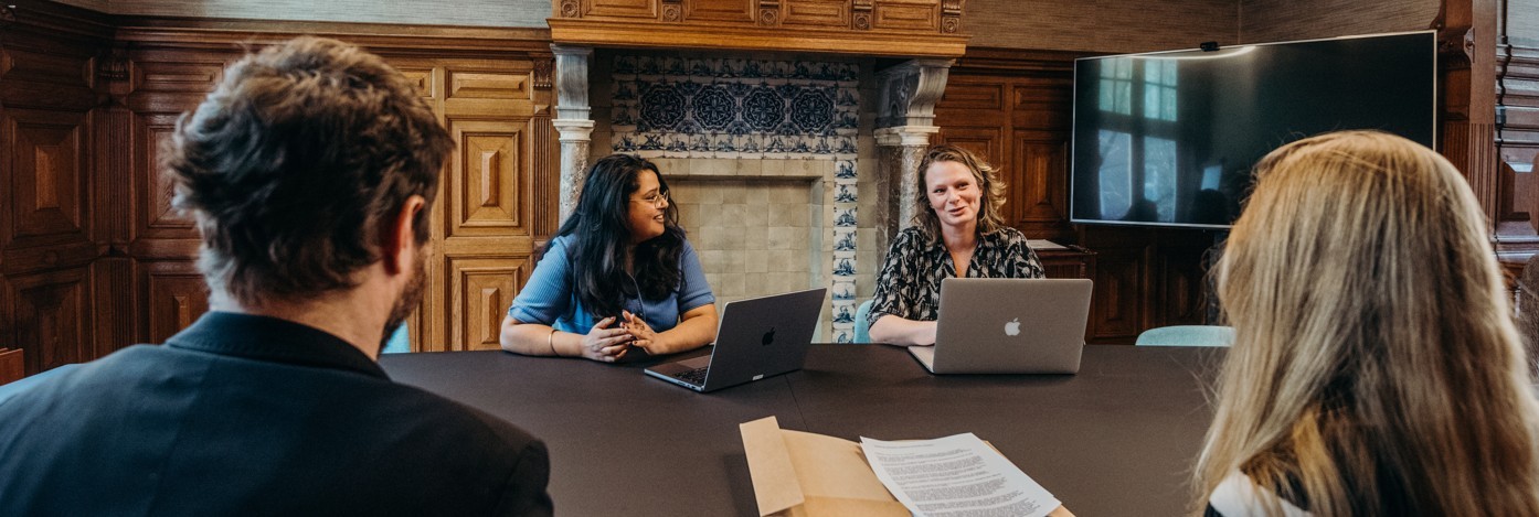 Twee vrouwelijke HuC-medewerkers zitten aan een tafel met hun laptops tegenover een man en een vrouw